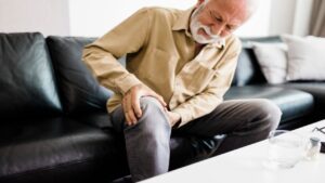 Senior Man Feeling Unwell, Suffering from Pain in the Leg While Sitting on a Sofa in the Living Room at Home