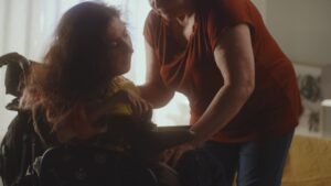 Aged Woman Caregiver Helping a Woman with Spinal Muscular Atrophy Sitting a Modern Motorized Wheelchair